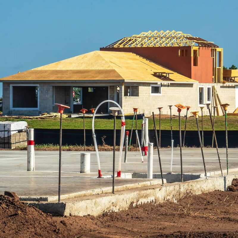 A residential construction site featuring a newly poured concrete slab foundation with exposed pipes and rebar in the foreground, and an unfinished house under a clear blue sky in the background.
