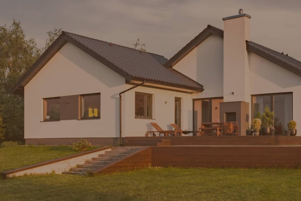 A modern white house with a dark roof and a spacious wooden deck sits on a lush green lawn under a warm sunset sky.