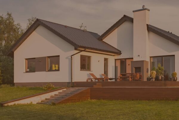 A modern white house with a dark roof and a spacious wooden deck sits on a lush green lawn under a warm sunset sky.