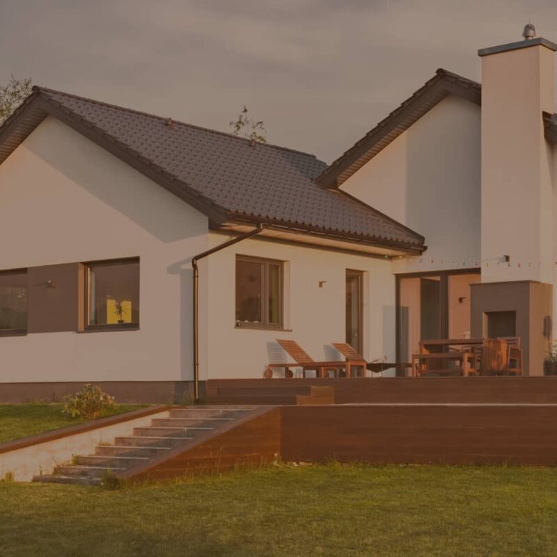 A modern white house with a dark roof and a spacious wooden deck sits on a lush green lawn under a warm sunset sky.