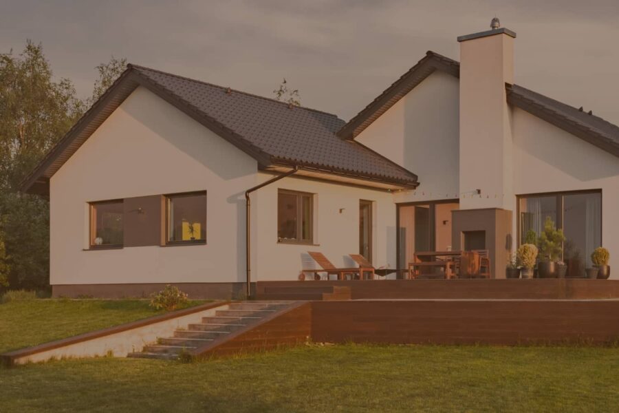 A modern white house with a dark roof and a spacious wooden deck sits on a lush green lawn under a warm sunset sky.