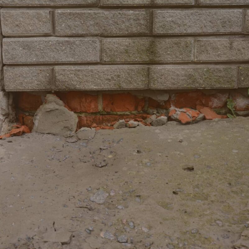A close-up view of a building's corner showing a severely damaged foundation with crumbling red bricks and broken masonry debris beneath a wall of grey blocks.