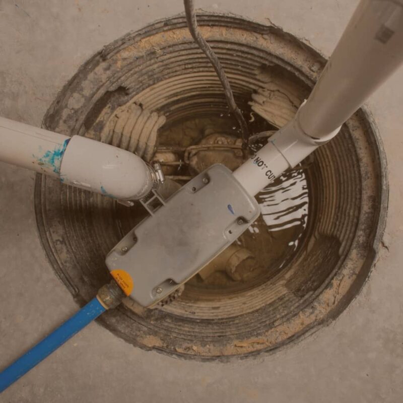 Top-down view of an open basement sump pump pit containing standing water, PVC discharge pipes, and electrical wiring.