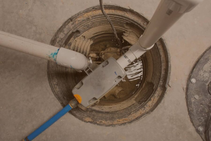 Top-down view of an open basement sump pump pit containing standing water, PVC discharge pipes, and electrical wiring.